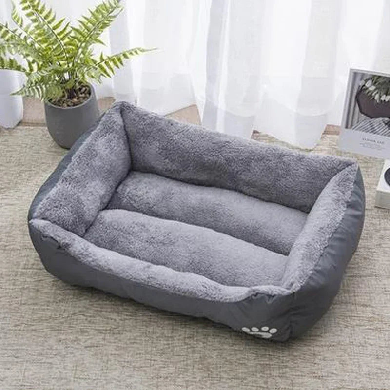 Plush rectangular gray pet bed with soft lining, placed on a carpet near a window and plant.